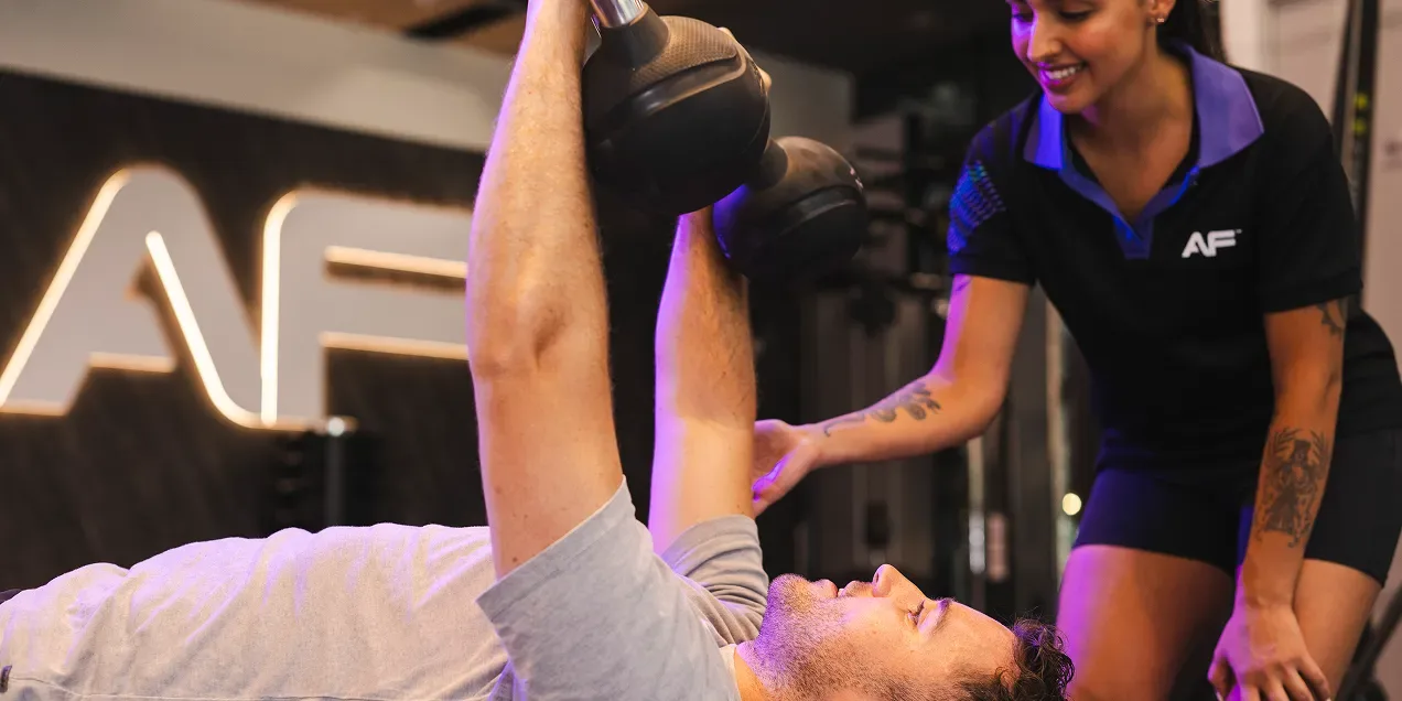 A man performs a bench press with dumbbells, while a trainer offers assistance in a modern gym setting.