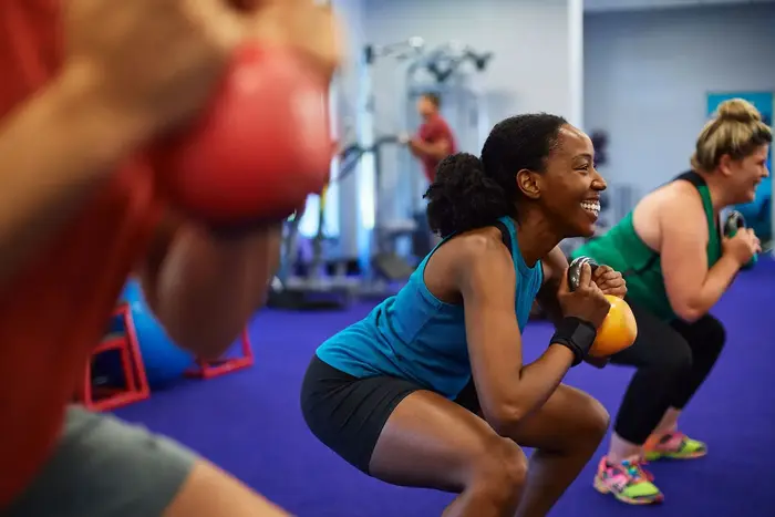 Three people in a gym doing squats while holding kettlebells, smiling and focused on exercise.